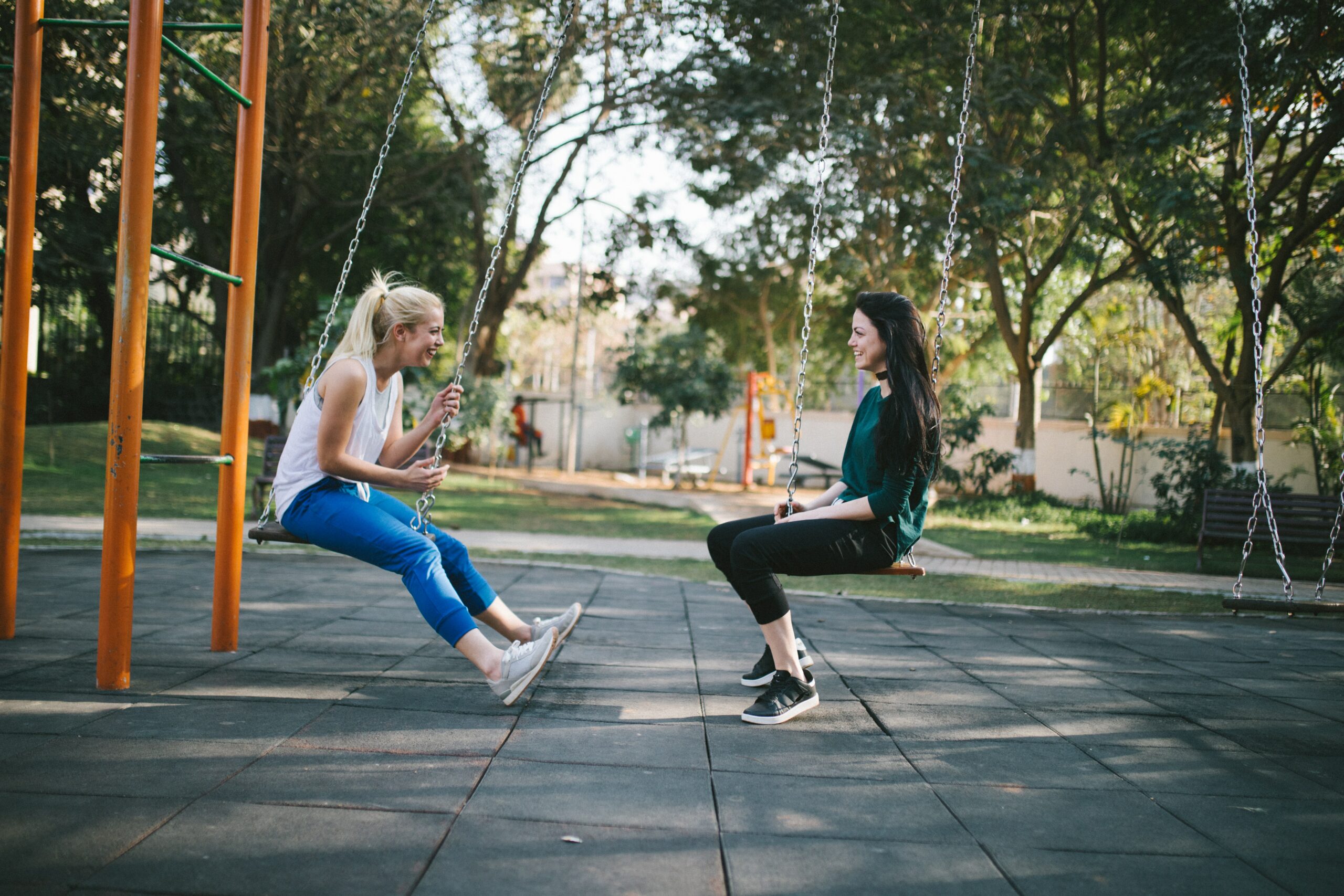 Two women sit and chat on a swingset in the park