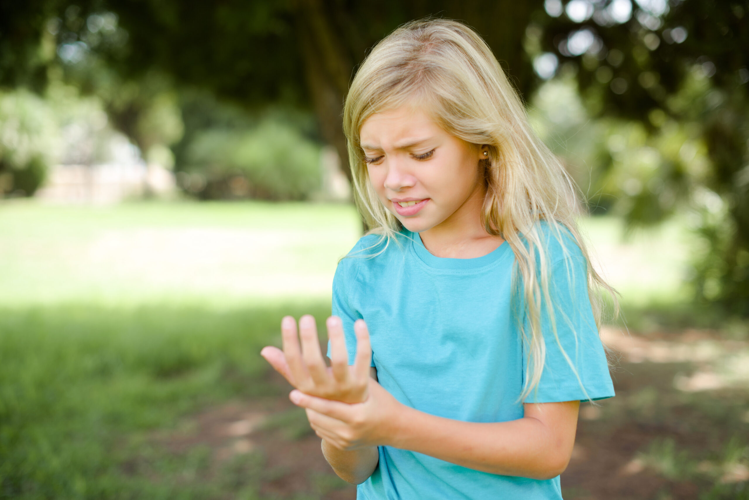 Caucasian little kid girl wearing blue T-shirt standing outdoors Suffering pain on hands and fingers, arthritis inflammation