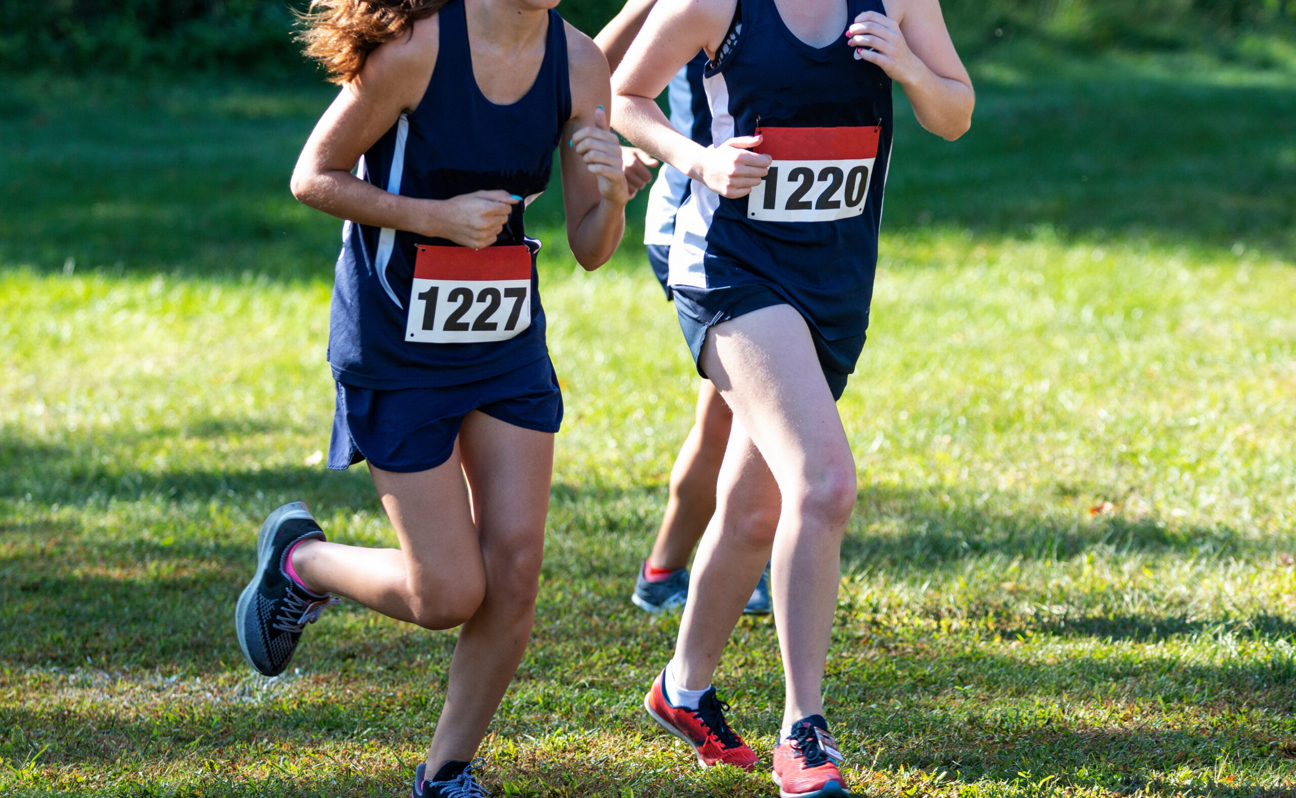 Three high school girls competing in a high school cross country race wearing blue uniforms on a grass field.
