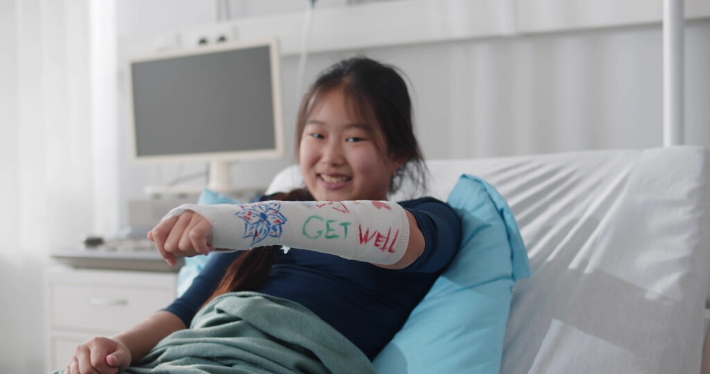 Smiling asian preteen girl lying in hospital bed with broken arm in colored cast