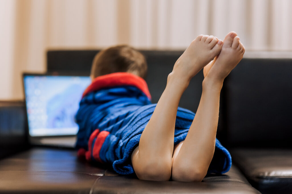 A boy child in pajamas lies on a black leather sofa in the evening, watching a movie on a laptop online via the Internet