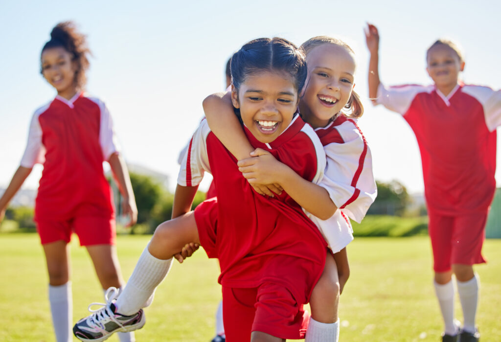 Soccer, girl celebration and field with happy piggyback, team building support and solidarity for winning game. Female kids, sports diversity and celebrate with friends, teamwork and goal in football.