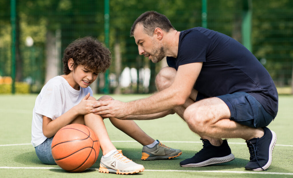 Side view portrait of focused adult man coach helping boy with knee trauma after playing basketball on the court, sad player feeling pain, touching leg. Activity And Sports Injury Concept.